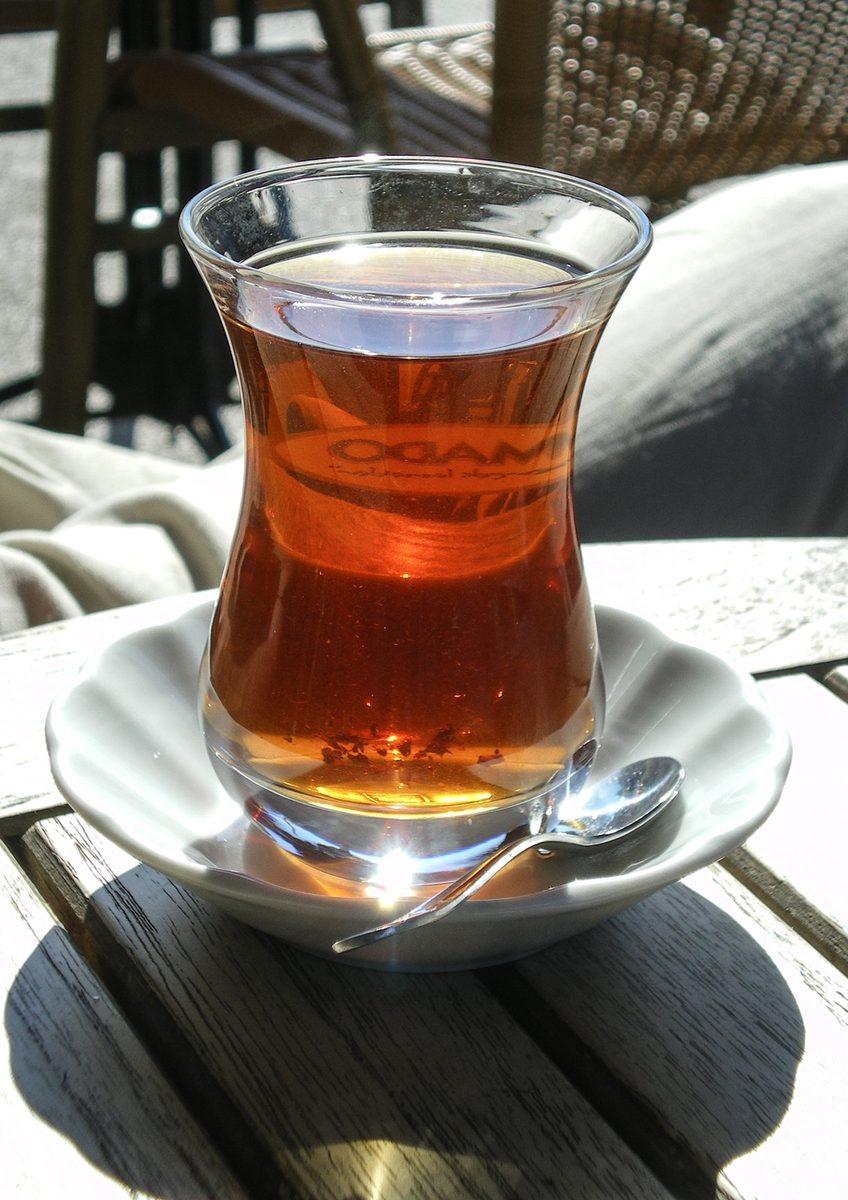 A tulip-shaped glass of Turkish tea on a white saucer with a spoon, in sunlight on a wooden table