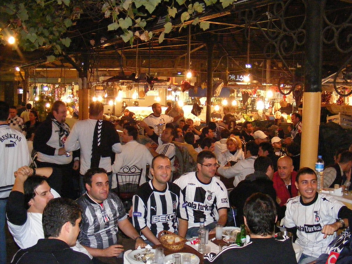 Beşiktaş football fans in black and white shirts eating at crowded tables under the fish market canopy in Besiktas Istanbul